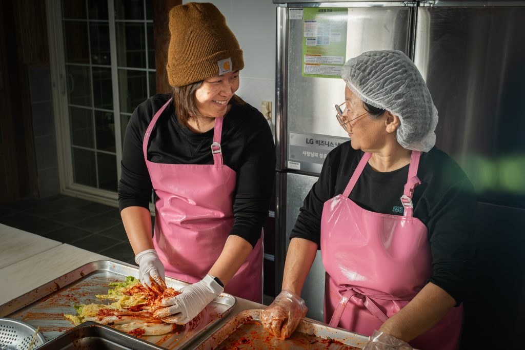 Chef Ji Hye Kim making kimchi with another person, both are wearing pink aprons